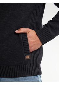 Mörk stickad tröja med ribbade mudd och nederkant, med en sidficka och en märke av läder nära nederkanten. Mjuk textur och avslappnad passform.