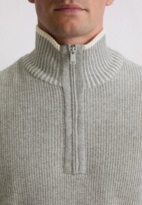 Pull-over gris à côtes avec un col haut et une fermeture éclair. Le col présente une bordure crème à côtes, soulignant la texture du tissu.