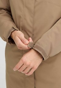 Beige gepolsterter Mantel mit Nahaufnahme des Handgelenkbereichs, der einen Klettverschluss und eine weiche Manschettenstruktur zeigt.