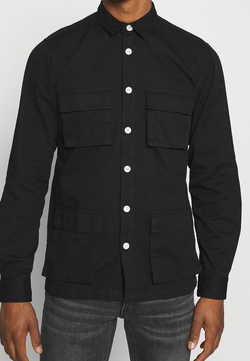 Homme portant une chemise noire à boutons avec quatre grandes poches avant et un jean gris foncé, debout les bras détendus.
