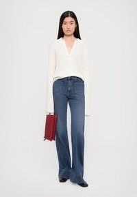 Top bianco a maniche lunghe a coste con scollatura a V, abbinato a jeans a zampa blu e scarpe nere, mentre tiene in mano una borsa rettangolare rossa.