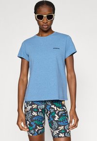 Γαλάζιο βαμβακερό μπλουζάκι με κοντά μανίκια και μικρό μαύρο λογότυπο. Συνδυασμένο με ποδηλατικά σορτς με σχέδια πολύχρωμων φύλλων.