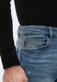 Svetlomodré džíny z denimu majú päťvreckový dizajn, jemné vyblednutie, efekty whiskering a štítok s logom na prednom vrecku. Nosené s čiernym svetrom.