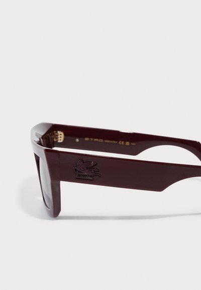 Lunettes de soleil bordeaux avec des montures rectangulaires épaisses, arborant un emblème en relief sur la branche, des verres teintés et une texture en plastique lisse.