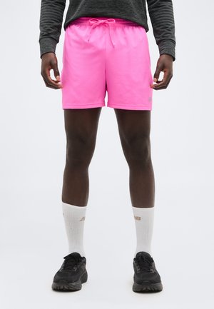 Mann som har på seg knallrosa shorts med snøring, hvite ankelsokker, svarte joggesko og en mørkegrå genser med lange ermer, står med armene avslappet.