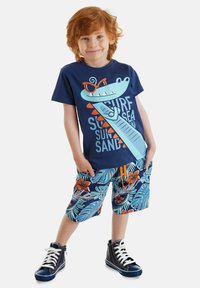 Marinefarbenes T-Shirt mit einem Cartoon-Krokodil-Design und Text. Bunte Shorts mit Blumenmuster und einer orangen Zugkordel. Schwarze High-Top-Sneaker.