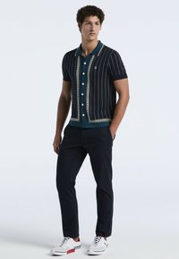 Jongeman staat rechtop, gekleed in een marineblauwe kortarmige button-up cardigan over een teal shirt, donkere broek en witte sneakers met rode accenten.