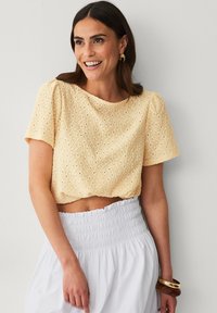 Lys gul, cropped top med hulmønstret og korte ærmer, parret med en hvid nederdel med smocket talje. Glat stofstruktur.