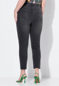 Vaqueros de mezclilla negra con una cintura alta, corte ajustado y costuras decorativas en los bolsillos traseros. Combinados con sandalias de tacón alto verdes.