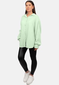 Camicia oversize verde chiaro con texture e bottoni, abbinata a leggings neri e sneakers bianche. Stile casual, stratificato.