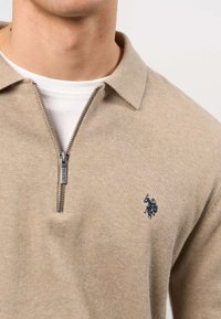 Man wearing beige zip-up sweater with collar over white shirt, featuring a small dark embroidered logo on the left chest.