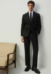 Musta puku hienovaraisella ruutukuviolla, valkoinen paita, yksivärinen musta solmio ja mustat kengät. Slim fit ja sileä texture, yhdistettynä vaaleanruskeaan tuoliin.