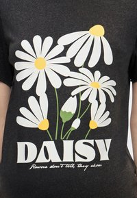 Μαύρο μπλουζάκι με λευκά και κίτρινα άνθη μαργαρίτας και μεγάλο κείμενο "DAISY" πάνω από μικρότερο κείμενο "Τα λουλούδια δεν λένε, δείχνουν."