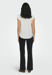 Top blanco sin mangas con detalles de volantes, combinado con pantalones negros de campana. La modelo está de espaldas frente a la cámara, luciendo zapatos marrones.