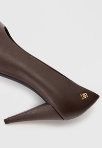 Brune lærhæl med spids tå og unik skrå hæl, der har et guld logo-accent på den ydre side.