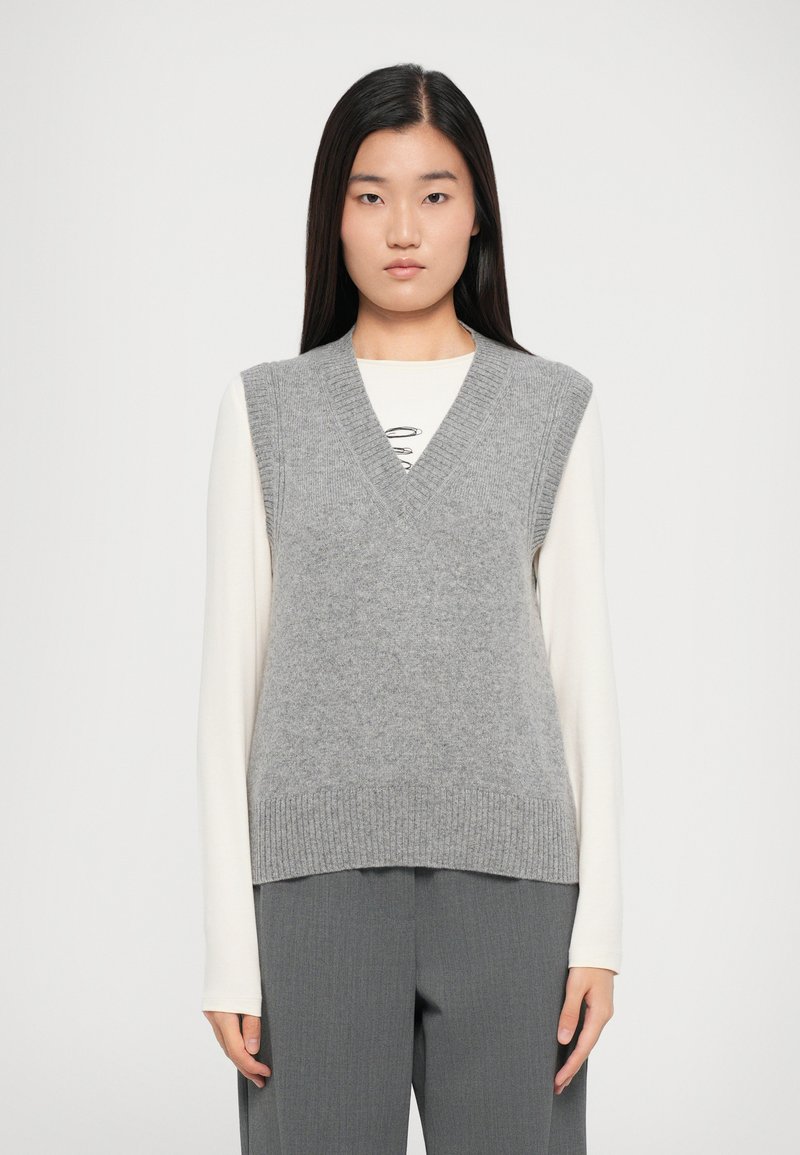 Gray knitted vest with a v-neck design, ribbed edges, and soft texture, worn over a long-sleeve cream shirt. Model stands front-facing.