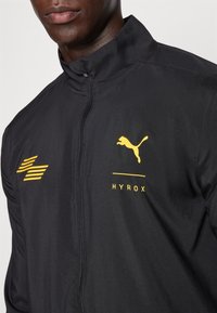 Chaqueta deportiva negra con cuello alto, cierre de cremallera, logotipo amarillo de Puma y texto "HYROX", hecha de material suave y ligero.