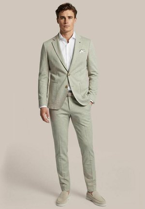 Man in lichtgrijs pak met wit overhemd en pochet, staand tegen een effen beige achtergrond met één hand in zijn zak.