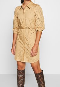 Vestido camisería color beige con mangas abullonadas, patrón de ojetes y un lazo en la cintura. Estilizado con botas altas de textura de serpiente.