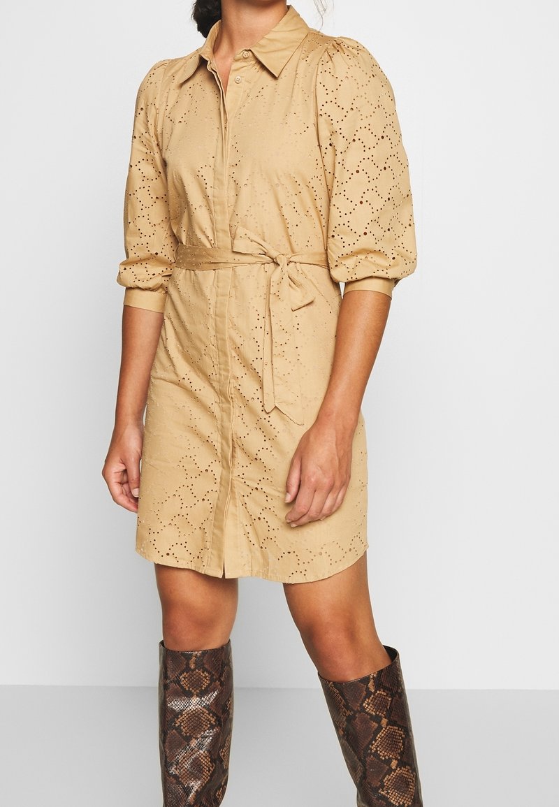 Vestido camisería color beige con mangas abullonadas, patrón de ojetes y un lazo en la cintura. Estilizado con botas altas de textura de serpiente.