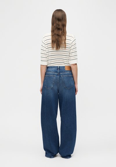 Femme aux longs cheveux portant un haut blanc à manches courtes rayé et un jean bleu à jambes larges, debout de dos sur un fond uni.