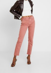 Kvinna som står i en croppad vit tröja, mörkbrun läderjacka, rosa högmidjade jeans och svarta högklackade sandaletter.