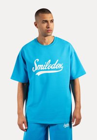 Camiseta oversized de algodón azul con un logo blanco de "Smilodox", cuello redondo y mangas cortas, acompañada de pantalones cortos azules a juego.