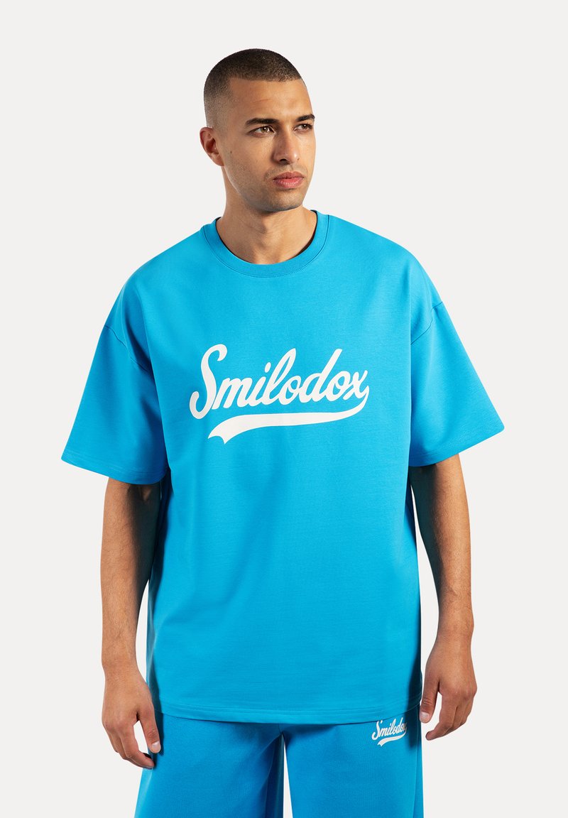 Camiseta oversized de algodón azul con un logo blanco de "Smilodox", cuello redondo y mangas cortas, acompañada de pantalones cortos azules a juego.