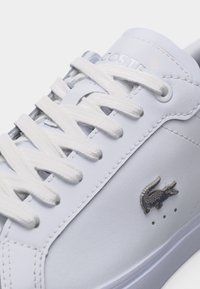 Λευκό δερμάτινο αθλητικό παπούτσι με λευκά κορδόνια, λογότυπο κροκόδειλου από ανοξείδωτο ατσάλι στο πλάι και ανάγλυφο όνομα της μάρκας στη γλώσσα.