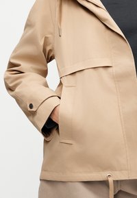 Veste beige avec poche à rabat et manchette à bouton-pression, portée avec la main dans la poche, sur un pantalon beige et un haut intérieur noir.
