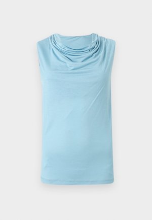 Γυναίκα που φοράει ένα αμάνικο ανοιχτό γαλάζιο τοπ με drape, μπέζ μίντι φούστα με μπροστινή σχισμή και μαύρα μυτερά παπούτσια, στέκεται μπροστά σε λευκό φόντο.