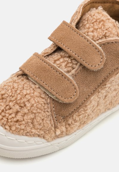 Tan sneaker with velcro straps, featuring a textured fuzzy material and suede accents. White rubber sole with stitch detailing.