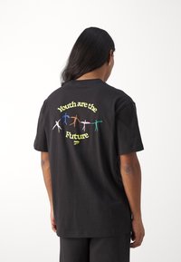 Osoba s dlouhými tmavými vlasy, oblečená v černém tričku se barevnými postavami a textem "Mladí jsou budoucností" na zádech, na bílém pozadí.