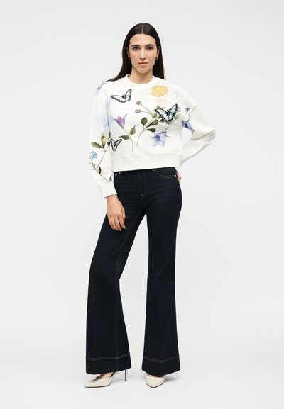 Γυναίκα όρθια με το χέρι στη μέση, φορώντας λευκό πουλόβερ με λουλουδάτα και πεταλούδες σχέδια, σκούρα φαρδιά τζιν και λευκά τακούνια.