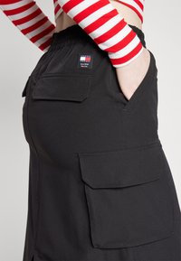 Sorte cargobukser med elastisk taljebånd, der har to store sidelommer og en label bagpå. Stoffet ser glat og struktureret ud.