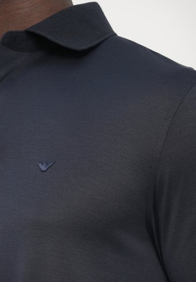 Tengerészkék galléros póló texturált gallérral, egy kis tónusos logóval a mellkasán. Simább anyag, testhez álló szabás.