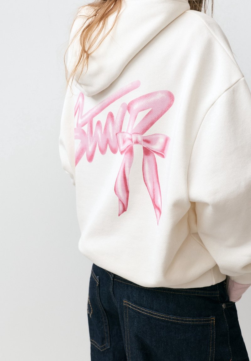 Personne portant un sweat à capuche blanc avec un texte style graffiti rose et un motif nœud dans le dos, associé à un jean bleu foncé.