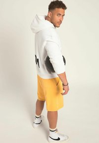 Vit huvtröja med svart tryck på ryggen, matchad med gula shorts. Tillbehör inkluderar en svart rund föremål och vita sneakers med svarta detaljer.