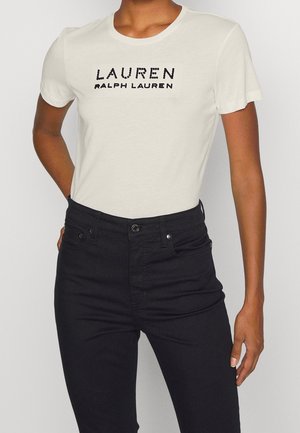 Osoba ubrana w białą koszulkę z krótkim rękawem z tekstem "Lauren Ralph Lauren" i czarne dżinsy z wysokim stanem na jednolitym tle.