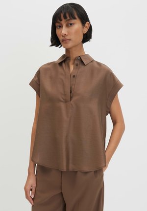 Frau mit kurzen schwarzen Haaren, die eine lockere braune Kurzarmbluse mit Kragen und passende braune Hose trägt, steht mit einer Hand in der Tasche.