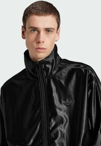 Svart zip-up jacka i glänsande material med hög krage, med en subtil tre-stripes design och en liten präglad logotyp på bröstet.