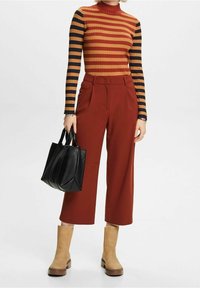 Femme portant un pantalon large couleur rouille, un col roulé à manches longues rayé, des bottes beiges, et tenant un sac fourre-tout en cuir noir.