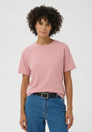 Frau mit lockigem Haar, die ein schlichtes rosa kurzärmeliges T-Shirt trägt, das in blaue Jeans mit einem schwarzen geflochtenen Gürtel gesteckt ist, vor weißem Hintergrund.