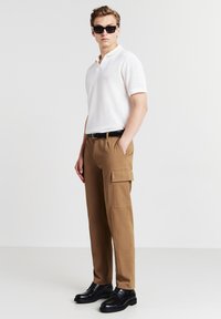 Polo shirt bianco a maniche corte in tessuto texture abbinato a pantaloni cargo beige con tasche laterali, indossato con scarpe nere stringate.