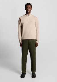 Felpa beige con zip e collo alto, maniche lunghe, abbinata a pantaloni dritti verde oliva e scarpe nere con suola spessa. Design semplice, senza motivi.