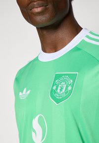 Zöld, rövid ujjú futballmez fehér díszítésekkel, a mellrészen Manchester United címer és Adidas logó látható. Sima anyagú textúra.
