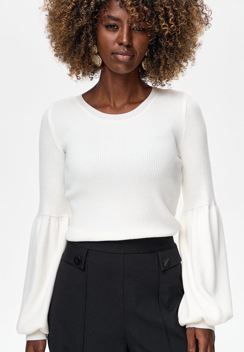 Vrouw met krullend haar, gouden oorbellen, een witte geribbelde trui met pofmouwen en een hoge taille zwarte broek met knoopdetails.