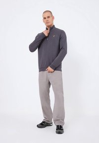 Grå hettegenser med glidelås og gul logodetalj, kombinert med lysegrå bukser og svarte sneakers. Jevn tekstur med en avslappet passform.