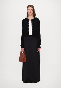 Cardigan nero con tasche frontali, bottoni e maniche lunghe indossato sopra una maglietta bianca. Gonna nera lunga fino al pavimento e borsa a manico marrone.
