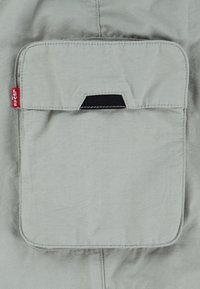 Poche en tissu gris clair avec rabat et détail en languette de tissu noir, étiquette rouge de la marque Levi’s sur le côté, cousue sur le vêtement.